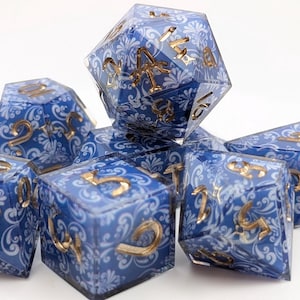 Könnte beinhalten: Ein Satz von sieben polyedrischen Würfeln. Die Würfel sind blau mit einem weißen Blumenmuster und goldfarbenen Zahlen. Die Würfel sind für Tabletop-Rollenspiele konzipiert.