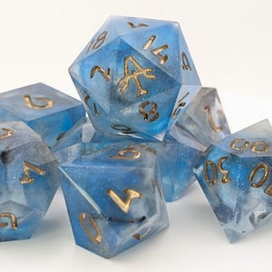 Könnte beinhalten: Ein Set aus sieben polyedrischen Würfeln in Blau mit goldenen Zahlen und Symbolen. Die Würfel haben ein himmlisches Design und sind für Tischrollenspiele gedacht. Die Würfel sind transparent.