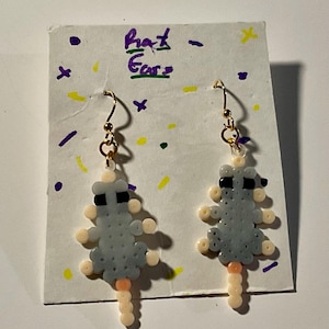 May include: A pair of handmade earrings featuring a gray rat design made from small beads. The earrings have gold-colored hooks and a light-colored tail. The earrings are displayed on a white card with the text "Rat Ears".