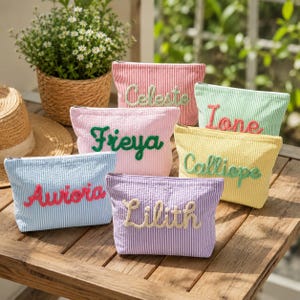 May include: A collection of six rectangular zippered pouches made of striped fabric in various pastel colors. Each pouch features a name embroidered in a contrasting color yarn on the front: Celeste, Ione, Freya, Calliope, Aurora, and Lilith.