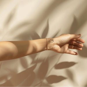 Peut inclure: Un bracelet dor&eacute; et un petit tatouage de fant&ocirc;me sur un poignet. Le tatouage est un contour noir d'un fant&ocirc;me avec un visage souriant. Le fond est un tissu beige avec des ombres.