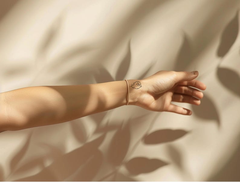Peut inclure: Un bracelet dor&eacute; et un petit tatouage de fant&ocirc;me sur un poignet. Le tatouage est un contour noir d'un fant&ocirc;me avec un visage souriant. Le fond est un tissu beige avec des ombres.