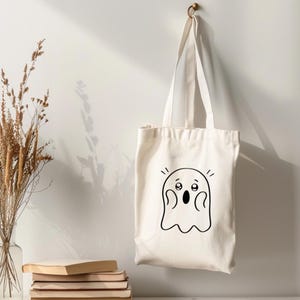 Peut inclure: Un sac fourre-tout cr&egrave;me avec un dessin de fant&ocirc;me de dessin anim&eacute; noir. Le fant&ocirc;me a de grands yeux et une bouche ouverte, lui donnant une expression surprise. Le sac est suspendu &agrave; un crochet en laiton sur un mur blanc.