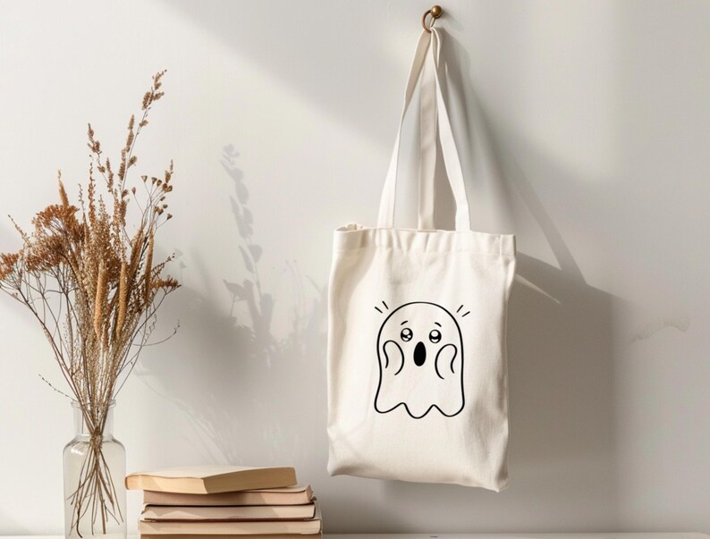 Peut inclure: Un sac fourre-tout cr&egrave;me avec un dessin de fant&ocirc;me de dessin anim&eacute; noir. Le fant&ocirc;me a de grands yeux et une bouche ouverte, lui donnant une expression surprise. Le sac est suspendu &agrave; un crochet en laiton sur un mur blanc.