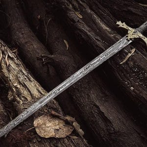May include: A long sword with a patterned blade and an ornate gold-coloured crossguard and hilt. The sword is displayed on a bed of dark brown wood, creating a contrast between the metal and the natural background.