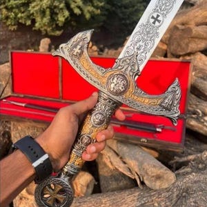 Puede incluir: Una espada ornamentada con una hoja plateada y detalles dorados. El mango presenta diseños intrincados y un pomo en forma de cruz. La espada se exhibe en una caja de madera con interior rojo, lo que sugiere un artículo de colección o decorativo.