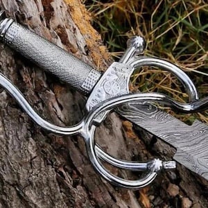 Pode incluir: Uma espada prateada com uma empunhadura texturizada e uma lâmina estampada. A espada apresenta uma guarda complexa com elementos curvos. A lâmina tem um padrão único em espiral. A espada está sobre um pedaço de madeira.