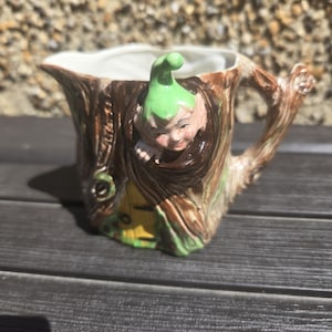 Op de afbeelding: Een keramische creamer in de vorm van een boomstam, met een handvat en tuit. Een kleine elf met een groene hoed en roze wangen kijkt uit een gat in de stam. De creamer heeft een witte binnenkant en is geschilderd in bruin, groen en geel.