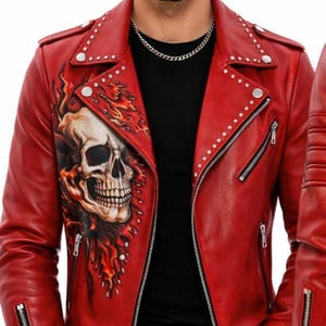 May include: Red leather jacket with a skull and flames graphic. The jacket has a classic biker style with silver zippers, studs, and a notched collar. The model is wearing a black shirt and black jeans.