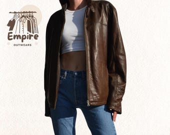 Puede incluir: Chaqueta de cuero marrón con cierre de cremallera. La chaqueta tiene un diseño clásico y se lleva abierta. La imagen también muestra un top blanco y vaqueros azules. El logo "Empire Outwears" es visible en la esquina superior izquierda.
