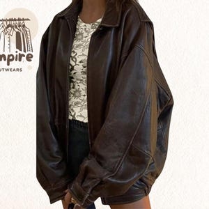 Pode incluir: Casaco de couro castanho escuro com fecho de correr e gola clássica. O casaco tem um corte folgado e um aspeto vintage. O logótipo da marca "Empire Outwears" é visível no canto superior esquerdo.