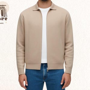 Pode incluir: Casaco bege com z&iacute;per e gola, usado sobre uma camiseta branca e jeans azuis. O casaco tem z&iacute;per completo e punhos canelados. O logotipo "Empire Outwears" &eacute; vis&iacute;vel no canto superior esquerdo.