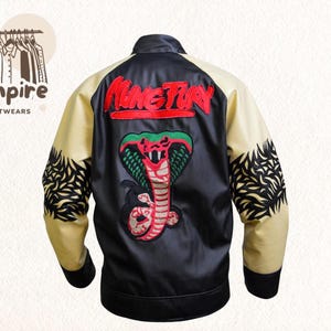 Puede incluir: Chaqueta de satén negro con mangas color crema y cuello negro. La espalda presenta un gran diseño de cobra bordada y la palabra "NINETEEN" en rojo. Las mangas tienen un patrón decorativo negro.