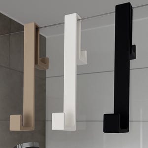 Op de afbeelding: Drie douchedeurhaken in beige, wit en zwart. Elke haak heeft een lange, rechthoekige vorm met een gebogen haak aan de onderkant en een bovenste gedeelte om over het glas te hangen. De haken hangen aan een glazen douchedeur.