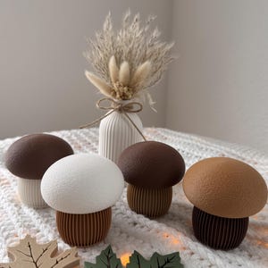 May include: A collection of decorative mushroom figurines with brown, white, and tan caps and ribbed brown bases. The mushrooms are arranged with wooden leaf cutouts and a small vase of dried flowers, creating a fall-themed display.