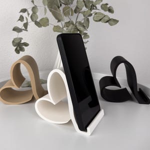May include: Three heart-shaped phone stands in beige, white, and black, with a smartphone in a black case. The stands are on a white surface, with a vase of greenery behind. The text "MACH3DRAUS" is visible.