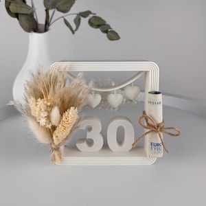 Könnte beinhalten: Ein weißer dekorativer Rahmen mit der Zahl "30" und drei herzförmigen Ornamenten. Ein kleiner Strauß Trockenblumen und eine aufgerollte Geldscheinrolle, die mit Bindfaden gebunden ist, sind ebenfalls vorhanden. Eine weiße Vase mit Eukalyptuszweigen befindet sich im Hintergrund.