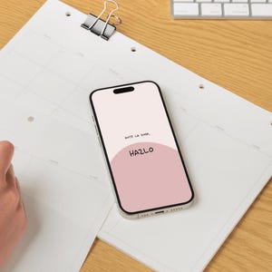以下が含まれることがあります： 「HAZLO」という単語が表示されたピンクと白の画面のスマートフォンが白いカレンダーの上に置かれています。手は黒いペンでカレンダーに書いています。コーヒーカップとキーボードも表示されています。