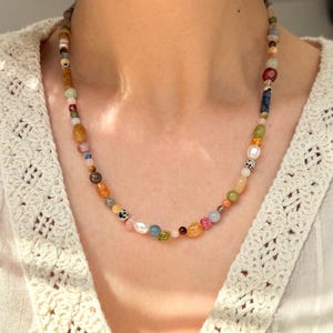 May include: A colorful beaded necklace with a variety of shapes and sizes. The necklace features a mix of earth-toned and pastel beads, including pearls, and is worn around the neck. The background is a white, textured top.