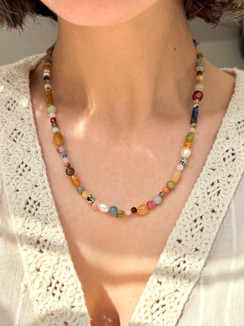 May include: A colorful beaded necklace with a variety of shapes and sizes. The necklace features a mix of earth-toned and pastel beads, including pearls, and is worn around the neck. The background is a white, textured top.