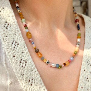 May include: A multi-colored beaded necklace with various shapes and sizes of beads. The necklace includes beads in shades of yellow, orange, pink, blue, green, and white. The necklace is worn over a cream-colored, open-knit sweater.