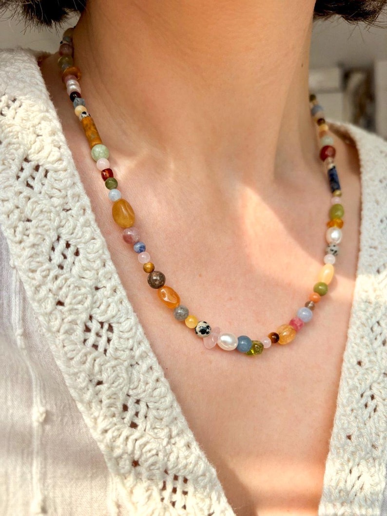 May include: A multi-colored beaded necklace with various shapes and sizes of beads. The necklace includes beads in shades of yellow, orange, pink, blue, green, and white. The necklace is worn over a cream-colored, open-knit sweater.