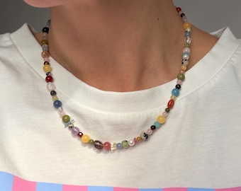 Peut inclure: Un collier de perles composé d'une variété de petites pierres et perles rondes et multicolores dans des tons de jaune, rouge, bleu, vert, rose et noir, entremêlées de perles transparentes et blanches, porté autour du cou sur un t-shirt rayé.