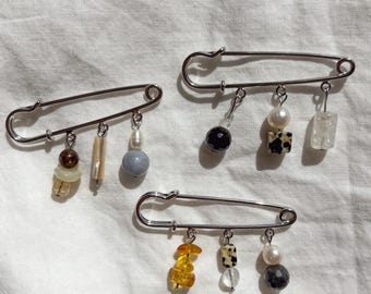 Peut inclure: Trois épingles à nourrice en métal argenté, chacune ornée de trois breloques pendantes. Les breloques sont composées de diverses perles, notamment des perles blanches, des cristaux translucides, des pierres ambrées et des perles tachetées.