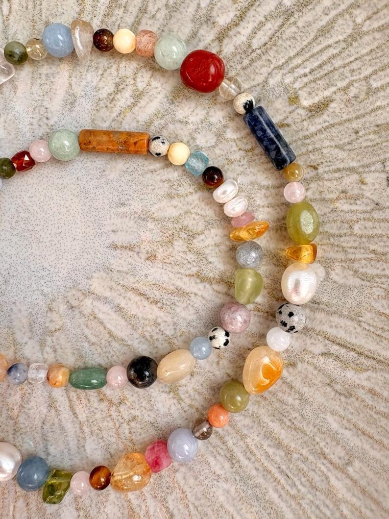 May include: A close-up of two beaded necklaces. The necklaces feature a variety of colorful gemstones and beads, including red, blue, green, and orange. The beads are strung together on a delicate string, creating a unique and eye-catching design.