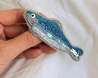 Peut inclure: Une broche en forme de poisson réalisée avec du fil à broder bleu et argent, ornée de sequins verts irisés sur le corps, d'une perle blanche pour l'œil et d'un contour en petites perles de rocaille argentées.