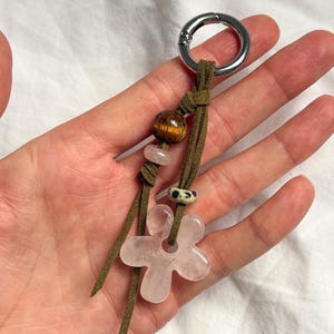 Op de afbeelding: Een sleutelhanger met een zilveren metalen ring en olijfgroene suède bandjes. De sleutelhanger heeft een houten kraal, een kleine roze steen, een patroonkraal en een roze bloemvormige stenen hanger. De sleutelhanger wordt in een hand gehouden.