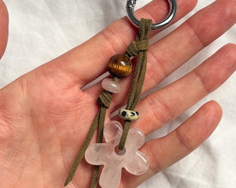 May include: A keychain with a silver metal ring and olive green suede straps. The keychain features a wooden bead, a small pink stone, a patterned bead, and a pink flower-shaped stone pendant. The keychain is held in a hand.