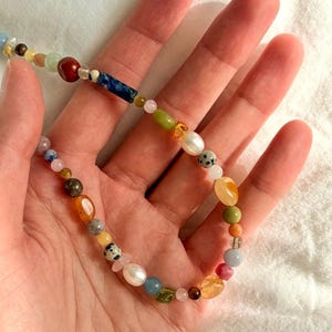 May include: A colorful beaded necklace featuring a variety of gemstones and pearls. The necklace includes beads in shades of red, blue, green, yellow, and white. The necklace is held in a hand.