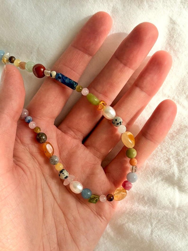 May include: A colorful beaded necklace featuring a variety of gemstones and pearls. The necklace includes beads in shades of red, blue, green, yellow, and white. The necklace is held in a hand.