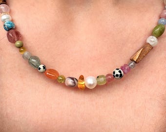 Peut inclure: Un collier de perles composé d'une variété de pierres précieuses multicolores, de perles et de perles à motifs. Le collier comprend des pierres lisses rondes, ovales et irrégulières dans des tons orange, vert, rose, bleu et blanc, ainsi qu'une perle rectangulaire brune texturée.
