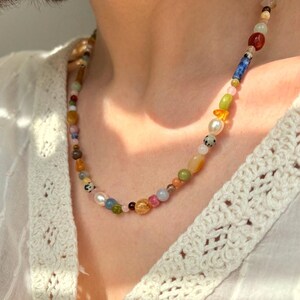 May include: A colorful beaded necklace featuring a variety of shapes and sizes. The necklace includes beads in shades of red, blue, green, yellow, and white. The necklace is worn over a white, crocheted top.