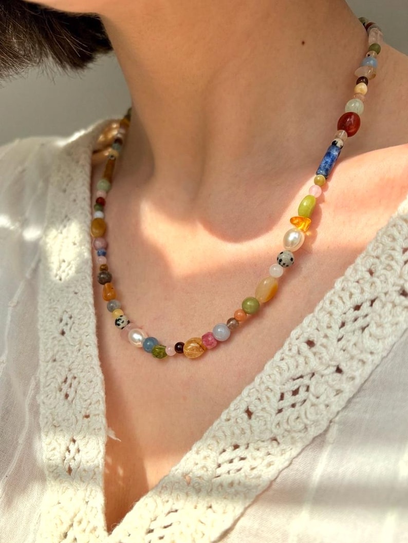 May include: A colorful beaded necklace featuring a variety of shapes and sizes. The necklace includes beads in shades of red, blue, green, yellow, and white. The necklace is worn over a white, crocheted top.
