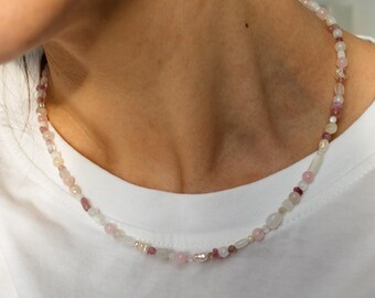 Peut inclure: Un collier délicat composé de diverses perles roses, blanches et transparentes. Les perles varient en taille et en forme, comprenant de petites perles rondes et des pierres plus grandes de forme irrégulière. Le collier est porté sur un t-shirt blanc.