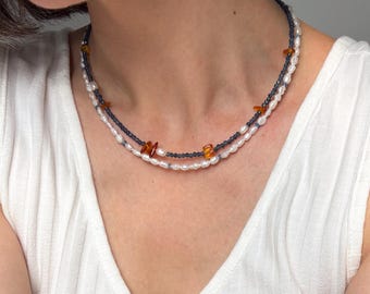Peut inclure: Un collier à double rang composé d'un rang de petites perles d'eau douce blanches et d'un second rang de perles gris foncé entrecoupées d'éclats de pierre irréguliers et translucides de couleur ambre autour du cou.