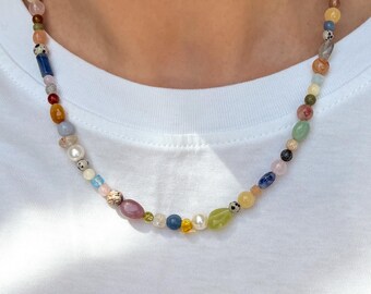 Peut inclure: Un collier de perles colorées avec une variété de pierres précieuses et de perles. Le collier présente un mélange de formes et de tailles, avec des teintes de bleu, rose, vert, jaune et marron. Le collier est porté sur un fond blanc.