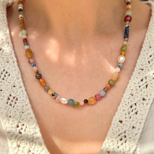 May include: A multi-colored beaded necklace featuring a variety of gemstone beads in shades of pink, yellow, green, and blue. The necklace includes a mix of round, oval, and irregular shaped beads, strung together on a delicate chain. The necklace is worn around the neck.