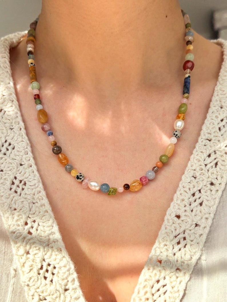 May include: A multi-colored beaded necklace featuring a variety of gemstone beads in shades of pink, yellow, green, and blue. The necklace includes a mix of round, oval, and irregular shaped beads, strung together on a delicate chain. The necklace is worn around the neck.