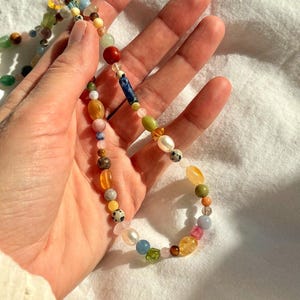 May include: A colorful beaded necklace featuring a variety of gemstones and beads in shades of red, blue, yellow, green, and pink. The necklace is held in a hand against a white background. The beads vary in size and shape.