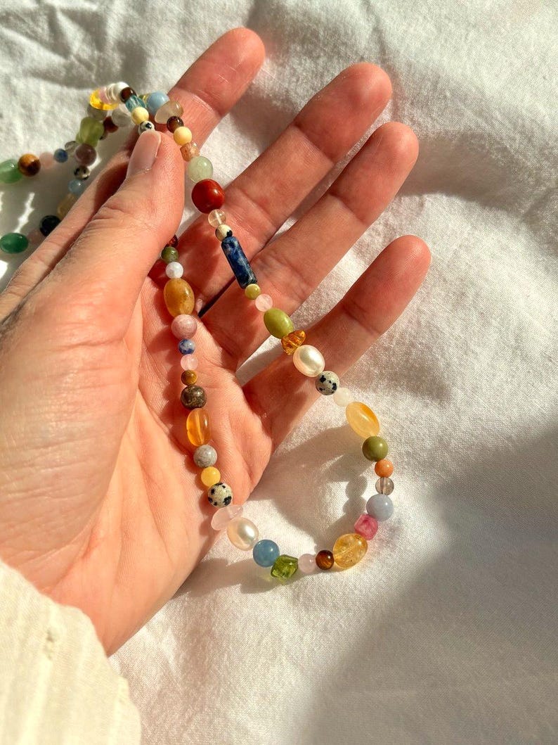 May include: A colorful beaded necklace featuring a variety of gemstones and beads in shades of red, blue, yellow, green, and pink. The necklace is held in a hand against a white background. The beads vary in size and shape.