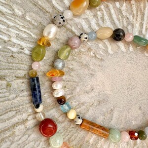 May include: A multi-strand necklace featuring a variety of colorful gemstones and pearls. The beads are in shades of orange, green, pink, blue, and brown. The necklace is arranged in a curved pattern, showcasing the different shapes and textures of the beads.