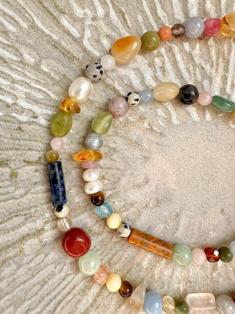 May include: A multi-strand necklace featuring a variety of colorful gemstones and pearls. The beads are in shades of orange, green, pink, blue, and brown. The necklace is arranged in a curved pattern, showcasing the different shapes and textures of the beads.