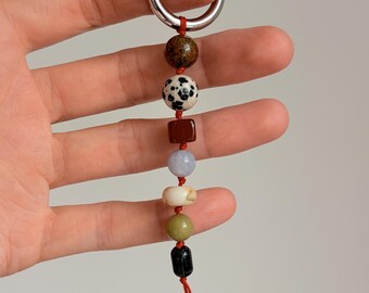 May include: A keychain featuring a silver-toned metal ring attached to a red cord. The cord is strung with seven beads in various shapes, sizes, and colors, including a brown speckled bead, a white bead with black spots, a red cube, and blue, white, green, and black beads.