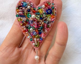 Peut inclure: Une broche en forme de cœur en feutrine rose, ornée d'une variété de perles en verre colorées, de paillettes et d'un cristal facetté central. Une perle blanche unique est suspendue à une épingle argentée au bas du cœur.