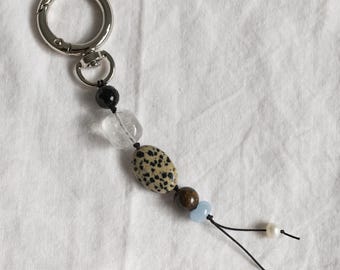 Peut inclure: Un porte-clés composé d'un anneau en métal argenté attaché à un cordon noir orné d'une perle noire à facettes, une perle transparente, une perle ovale en jaspe dalmatien, une perle brune, une perle bleu clair et deux petites perles blanches.