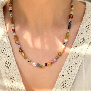 May include: A colorful beaded necklace featuring a variety of natural stone beads in shades of brown, green, yellow, pink, and blue. The necklace is worn around the neck and is a unique piece of jewelry.